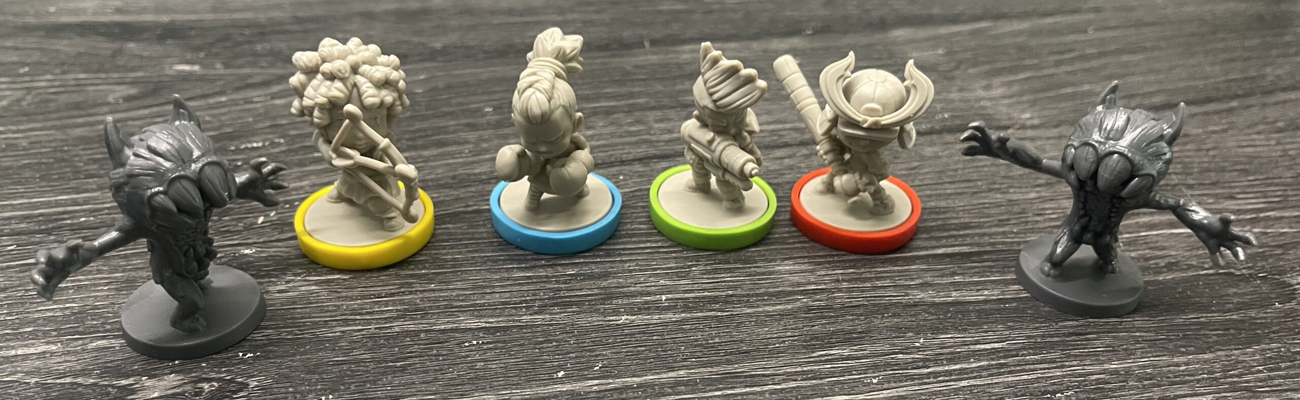 Row of seven small fantasy miniatures on a wooden surface: two dark, claw-handed monsters at the ends and five unpainted figures with various weapons in the middle, each on colored bases (yellow, blue, green, red).