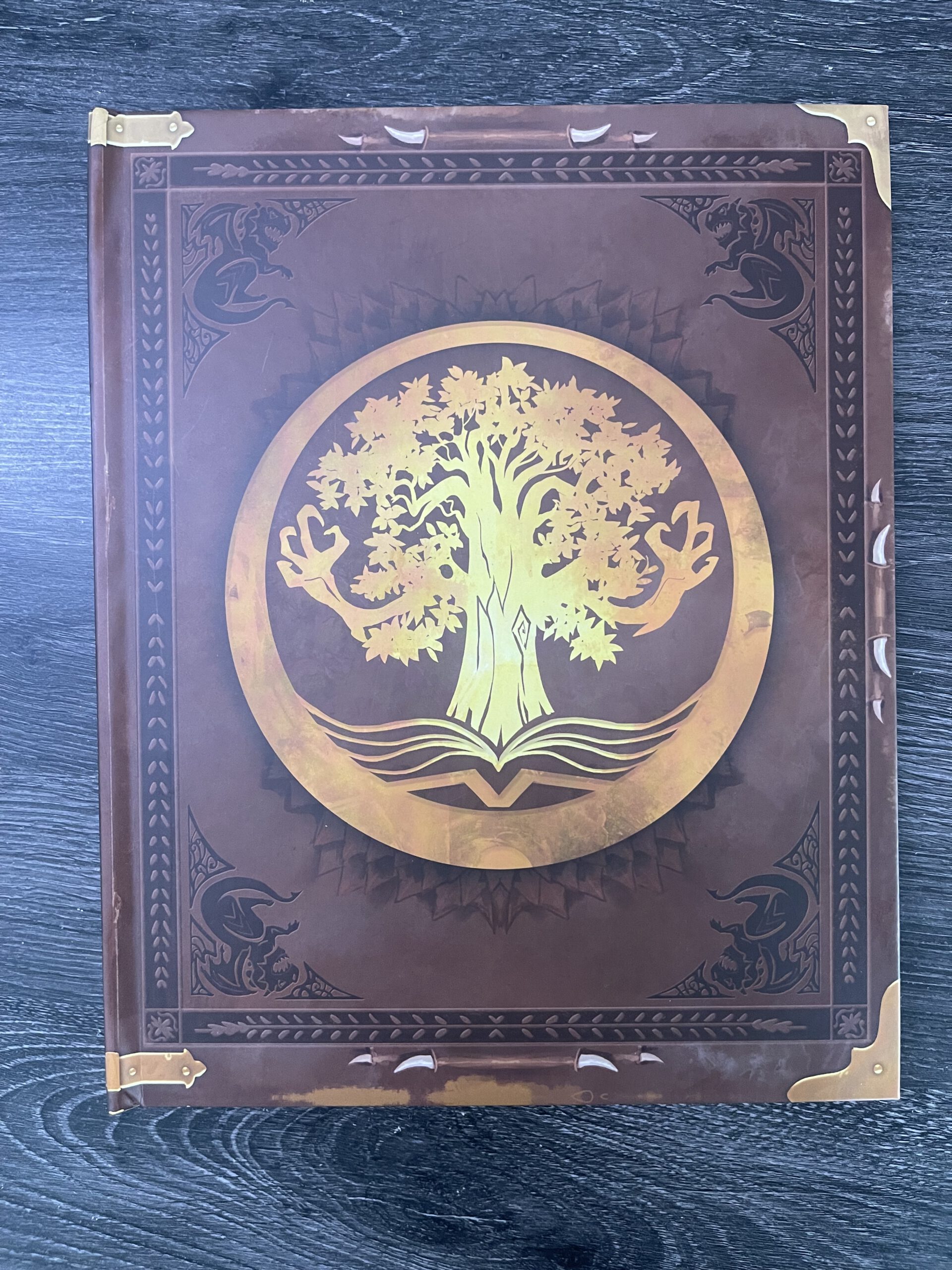Brown ornate book cover with a gold circular emblem of a tree, brass corner accents, and decorative borders -- a fantasy tome look.