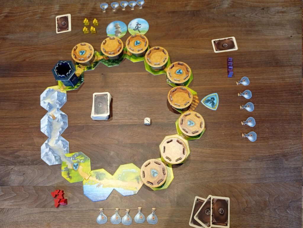Board game setup on a wooden table with modular hex tiles, round wooden town pieces, cards, dice, and small tokens ready for play.